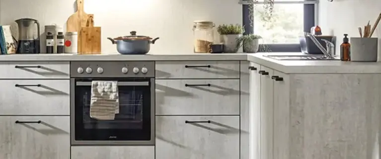 Einbauküche mit hellgrauen Unterschränken in L-Form, mittig Backofen mit Kochfeld und Topf, rechts Spüle am Fenster mit Pflanzen, auf der Arbeitsplatte Küchenutensilien und Gläser.